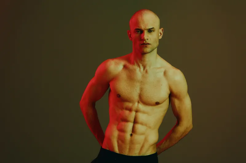 Strong man posing confidently, showing toned physique in a dramatic light setting. The composition highlights muscularity and determination in a captivating visual representation.