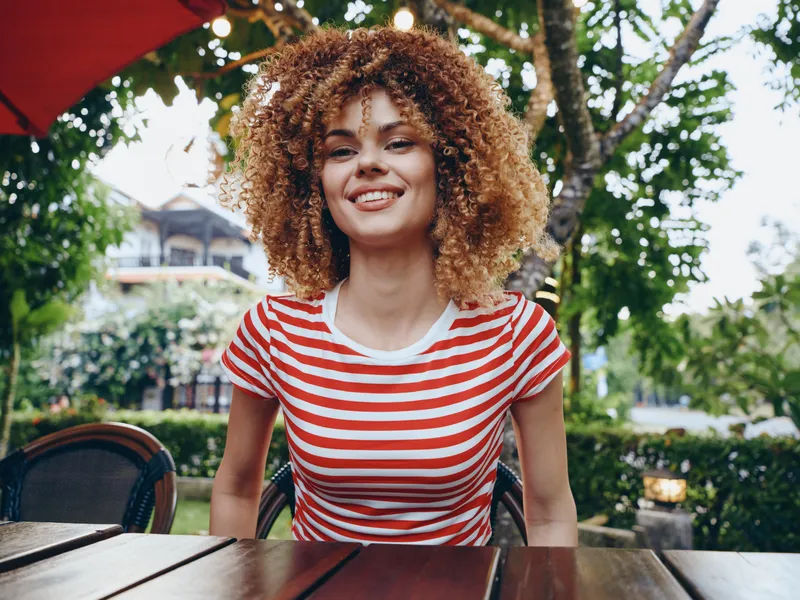 Young woman with curly hair sits at an outdoor cafe table, smiling warmly. She wears a red and white striped shirt, natural light, relaxed and welcoming vibe.