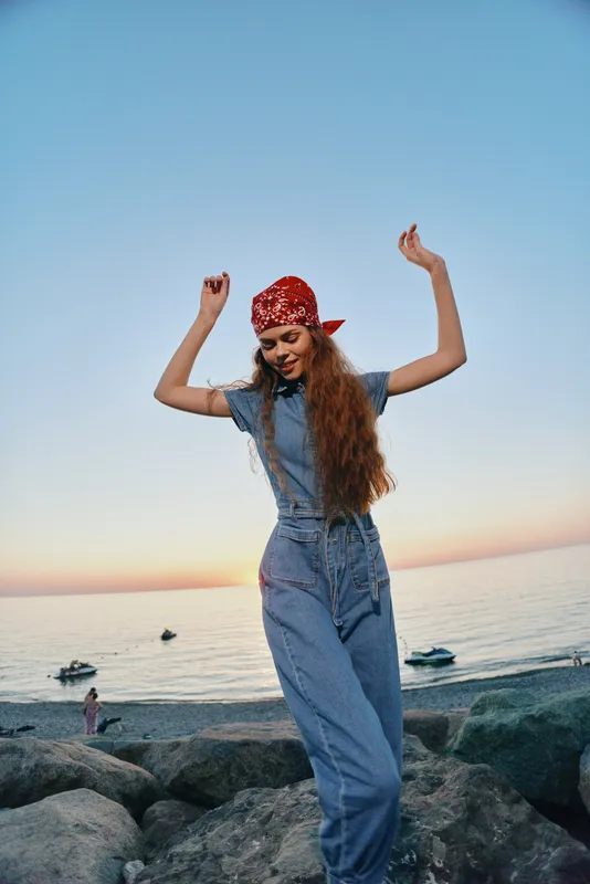 Lifestyle woman in modern boho-western denim street style dancing joyfully by the beach at sunset with film color tones and relaxed summer vibes.