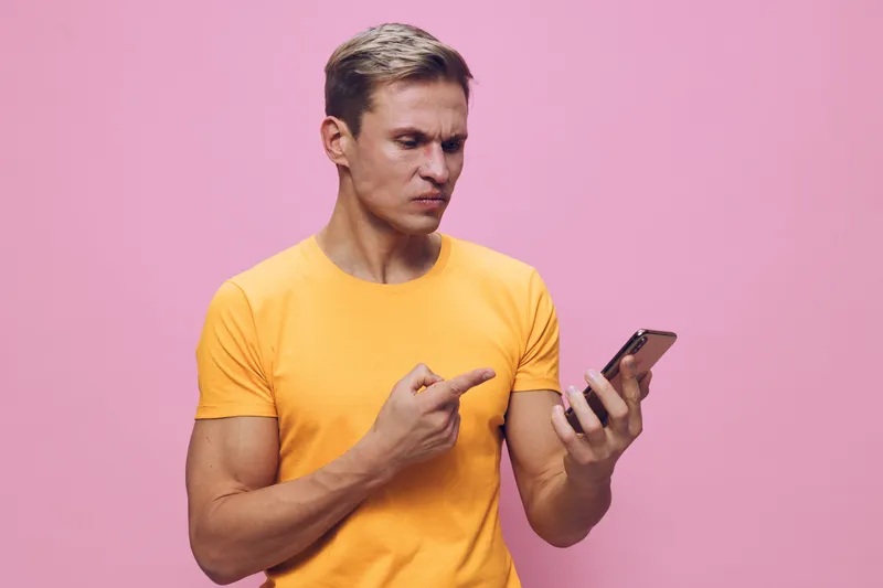 Man looks at smartphone with focused expression, points at screen, wears yellow t-shirt, pink background, casual style, technology, communication concept, young adult male, isolated on plain backgroun