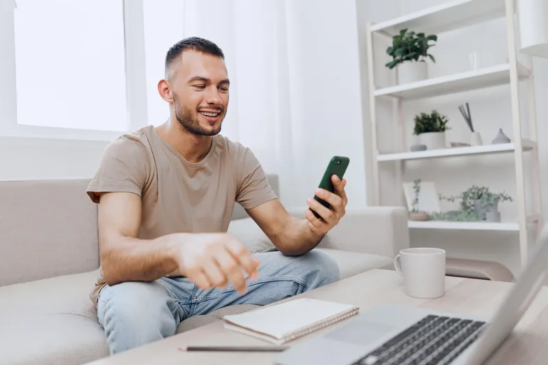 Smiling man enjoying his smartphone while sitting at home, surrounded by minimalistic decor and natural light. A relaxed atmosphere that reflects contemporary lifestyle choices.