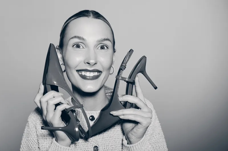 Smiling white woman with fair skin holding red high heels near face, wearing light sweater, isolated on grey background. Concept of fashion, shoes, style and happiness.