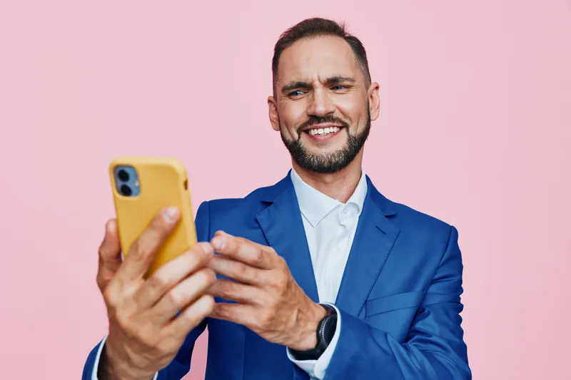 A professional man stands against a solid colored background, focused and confident while using a smartphone; an isolated scene that conveys ambition and a positive mood.