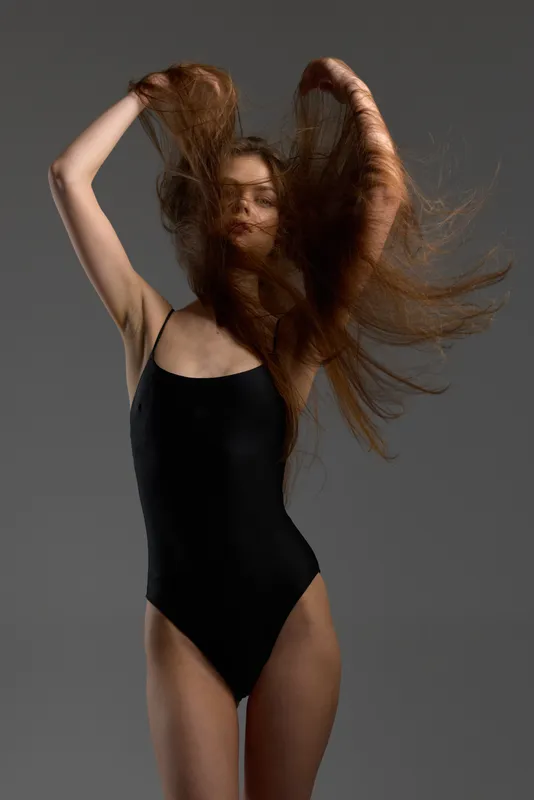 Young woman in black bodysuit with flowing hair on gray background expressing motion and freedom.