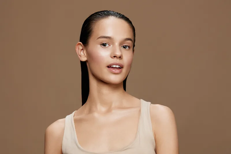 Young model with natural makeup and sleek hair, showcasing confidence and beauty against a warm brown background, ideal for beauty and skincare concepts