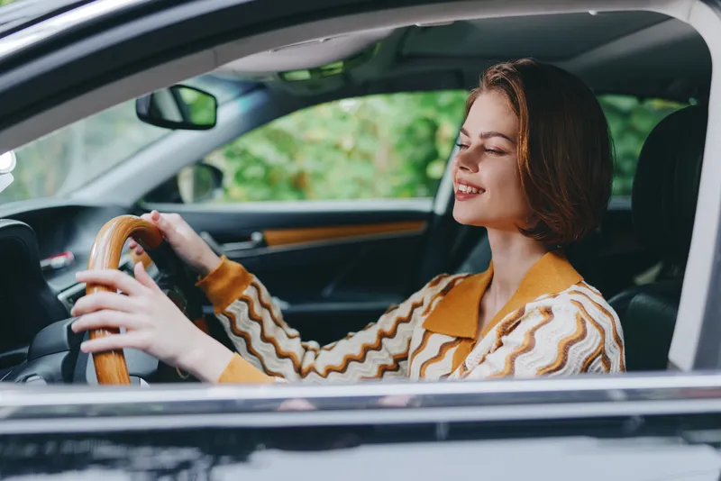 Happy woman driving a car with a wooden steering wheel, smiling as she enjoys a sunny day behind the wheel.