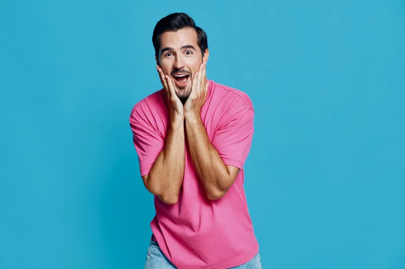Full body surprised cheerful young man wear pink t-shirt look camera with hands on face isolated on plain blue background studio portrait. Joyful expression concept