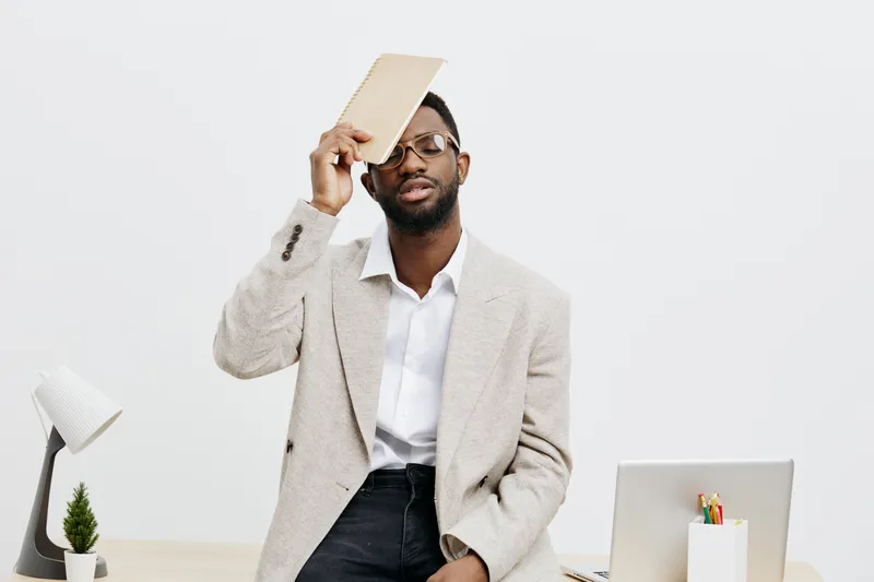 Professional stylish man holding book to his head while sitting at modern office desk full of supplies and tools, casual business outfit, thoughtful expression, indoor workspace, minimalist decor, nat