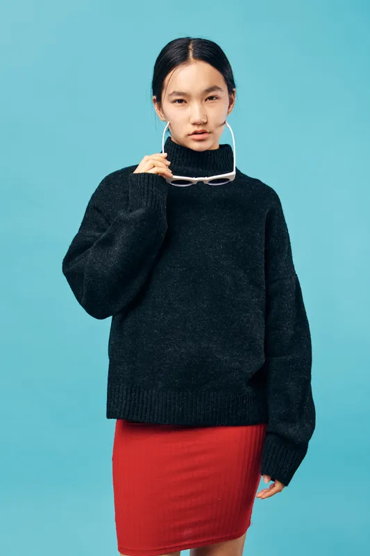 Young woman wearing black turtleneck sweater and red skirt posing on blue background. Female model with natural makeup and hoop earrings holding white sunglasses in studio.