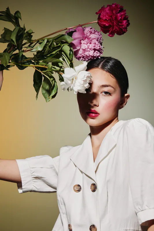 Stylish young woman in white blazer holding vibrant pink and white peonies above her face. Strong makeup with blush and red lips highlights beauty and fashion aesthetics.
