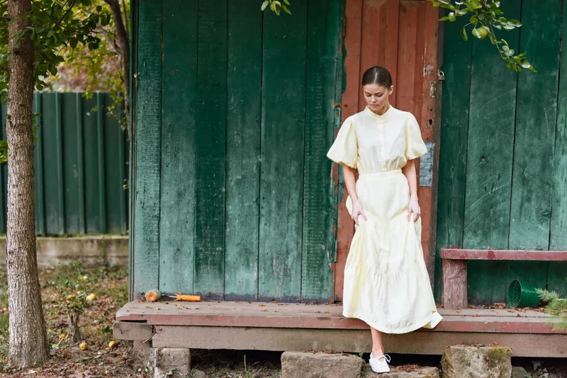 Young woman in a pale yellow dress standing on a wooden porch in a rustic outdoor setting, calm pose and soft light suggesting relaxed fashion moments
