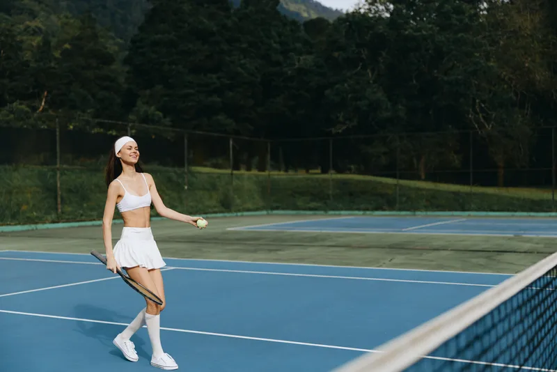Young woman enjoying a sunny day playing tennis in a white sports outfit on a court surrounded by green trees, expressing joy and vitality