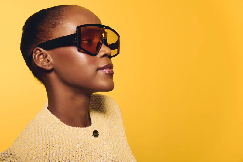 Profile of elegant black woman wearing large sunglasses and knitted sweater against yellow background. Concept of fashion, style, and confident female model with dark skin and afro hairstyle.