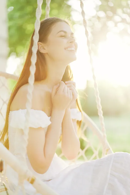 Joyful young woman in a white dress sitting on a swing, enjoying a sunny day in a lush green garden, radiating happiness and serenity