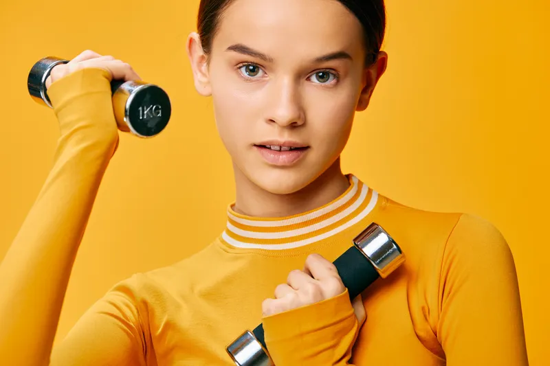 Young woman fitness portrait lifting small dumbbells in bright yellow sports top against vivid yellow background, focused expression and athletic posture conveying health and strength.