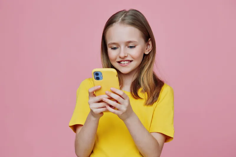 Smiling girl in a yellow shirt using a smartphone against a pink background, conveying joy and engagement with technology