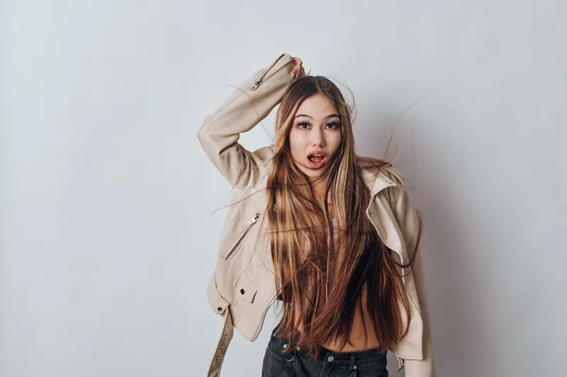 Young woman with long hair and casual beige jacket poses surprised against white background for fashion, style, and lifestyle concept with natural expression and modern outfit.