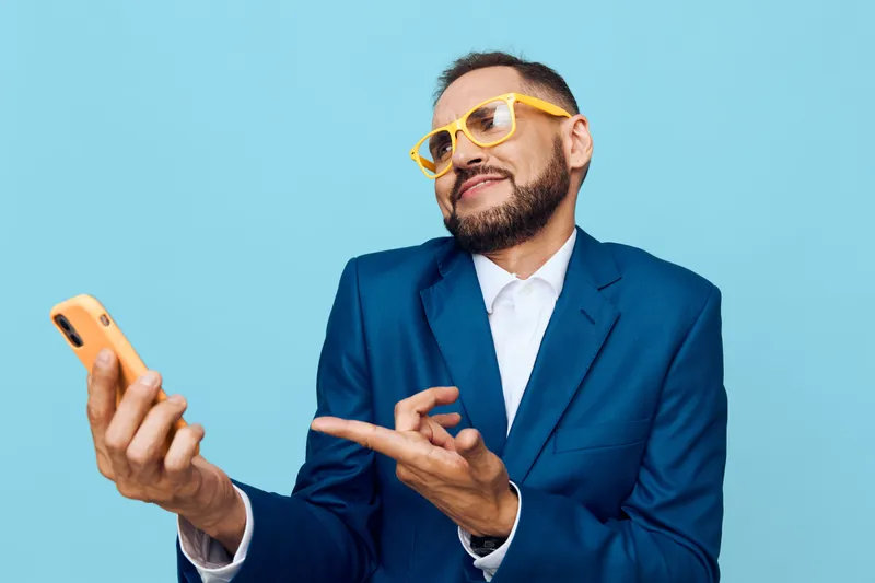 A professional man in a blue suit on a solid colored background, projecting calm confidence and curiosity, as he holds a smartphone and gestures with a subtle smile.