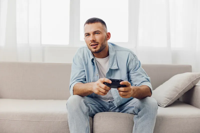 Engaged man playing video games on a cozy sofa, showcasing a modern lifestyle. His relaxed attire and focused expression reflect leisure and entertainment.