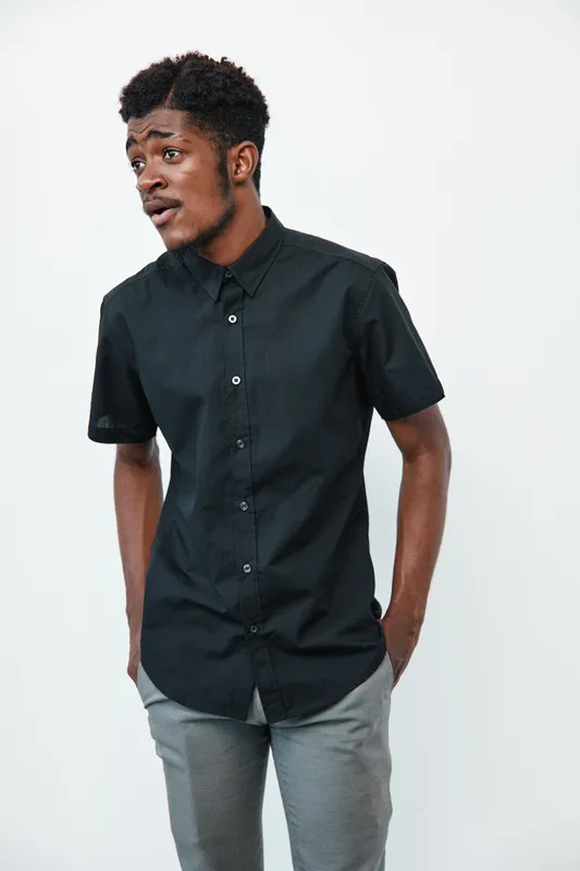 Young African man with dark skin wearing black short sleeve shirt and gray pants standing with hands in pockets on white background. Casual style and thoughtful expression.