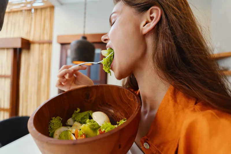Молодая женщина откусывает кусочек свежего салата на современной кухне