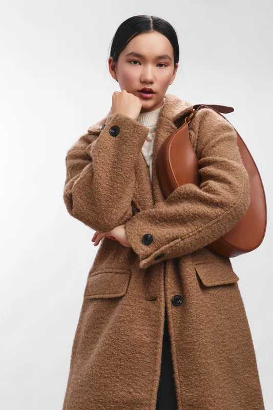 Asian woman confidently models in a textured brown coat and backpack, creating a chic streetwear look in a clean studio setting. Her poised expression and relaxed posture convey style and poise,