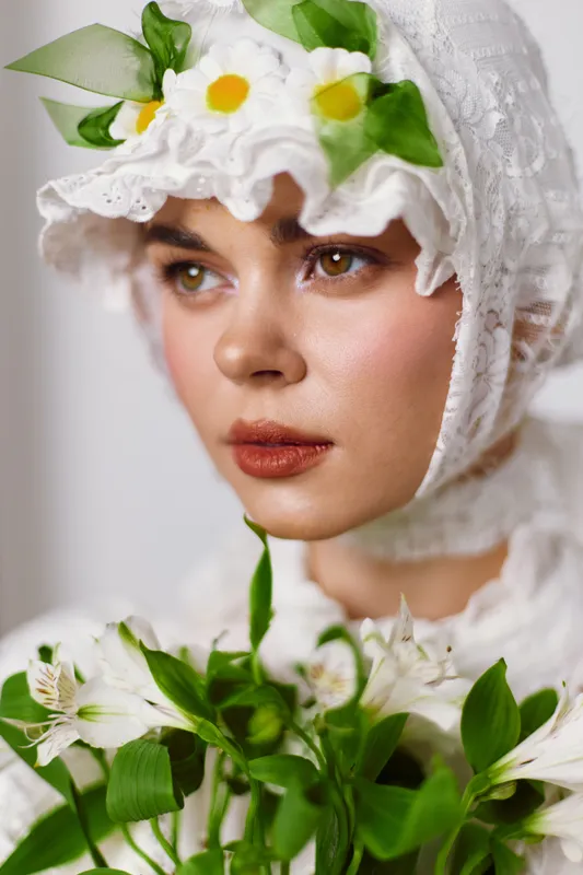 Young woman in traditional floral attire, featuring a white lace bonnet and light makeup, holding fresh flowers with a serene expression