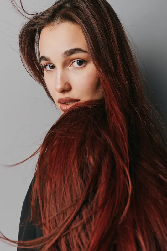 Woman with long red hair looking over shoulder with serious expression against plain background. Portrait of young female with natural makeup and flowing hair in studio lighting.