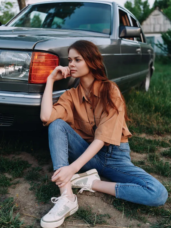 Fashionable young woman in casual outfit sitting next to classic car in summer, showcasing relaxed vibe and stylish look.
