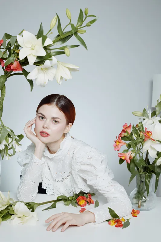 Young woman in elegant white blouse surrounded by fresh flowers, exuding serenity and beauty in a soft pastel setting