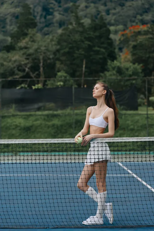 Young female tennis player in a white sports outfit exuding confidence on a tennis court, holding a tennis ball, set against a lush green background