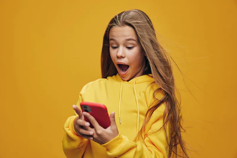 Girl with long hair wearing yellow hoodie looking surprised at red smartphone. Studio portrait on orange background with emotional expression and casual clothing for youth culture.