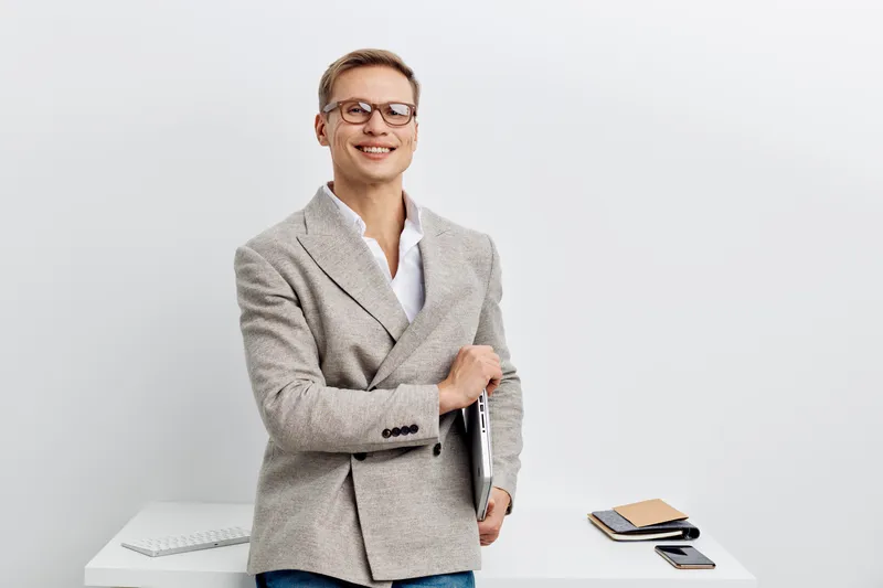 Smiling young Caucasian man in casual business attire standing in modern office with white background, holding a notebook and looking at camera, professional and friendly vibe, office lifestyle concep