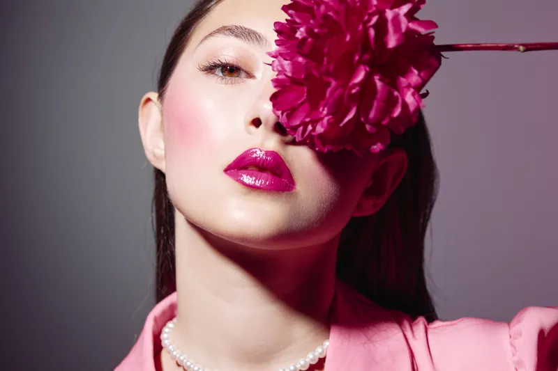 Close-up of a fashion model with glossy pink lips and shiny skin holding a vibrant magenta flower partially covering her eye. Contemporary beauty concept with creative fashion and elegance.