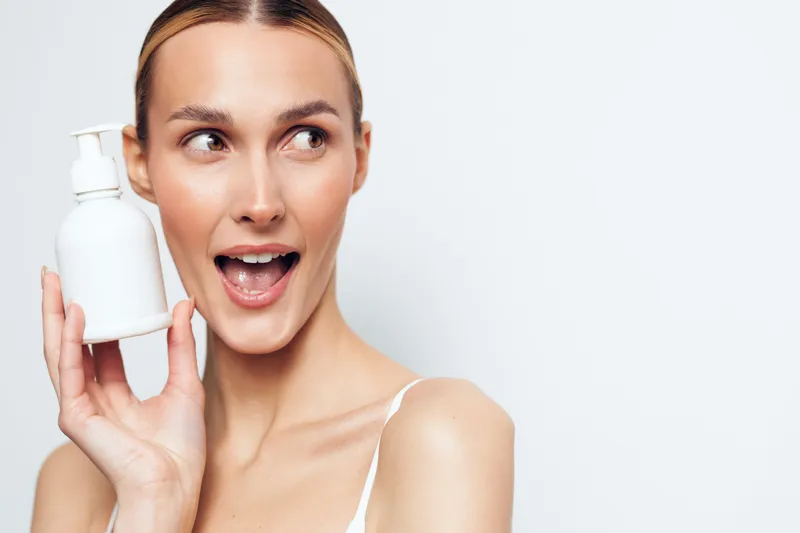 Young woman with clear skin holding a white lotion bottle near her face smiling with excitement on a plain background. Concept of skincare, beauty, and personal care.