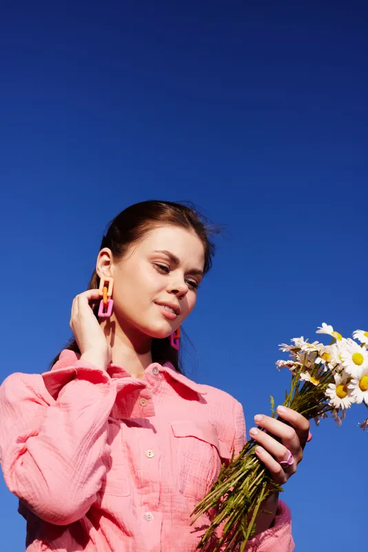 Young woman in a pink shirt holding daisies against a bright blue sky, expressing joy and appreciation for nature s beauty, showcasing spring vibes and floral themes