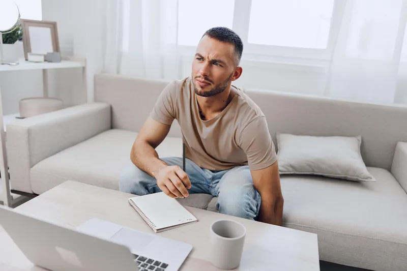 Thoughtful man engaging in remote work, surrounded by a modern home office atmosphere, contemplating ideas with a notepad, focused and inspired by creativity.