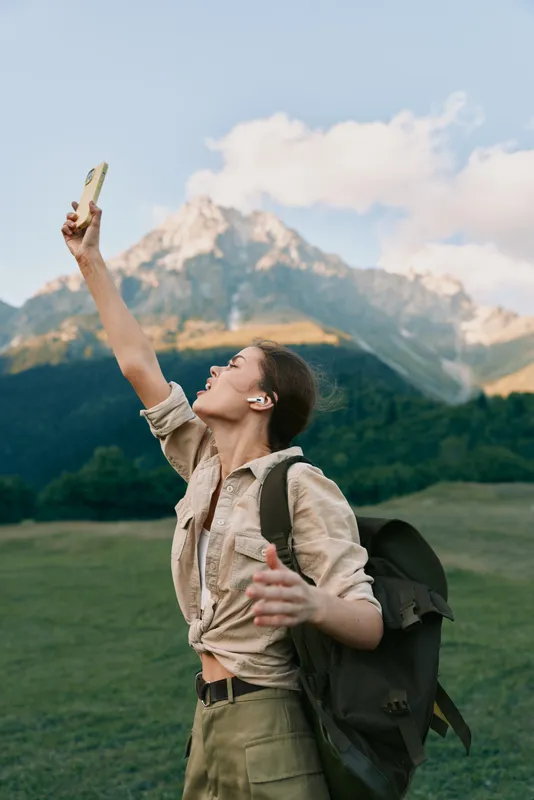 Young man with backpack raises his phone outdoors as towering mountains rise behind him, capturing a carefree selfie in a sunlit meadow.