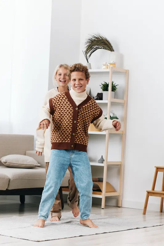 Two children dancing playfully in a bright living room, sunlight streaming in. Casual outfits, knitted vest, denim jeans, barefoot on a soft rug, capturing joyful movement.