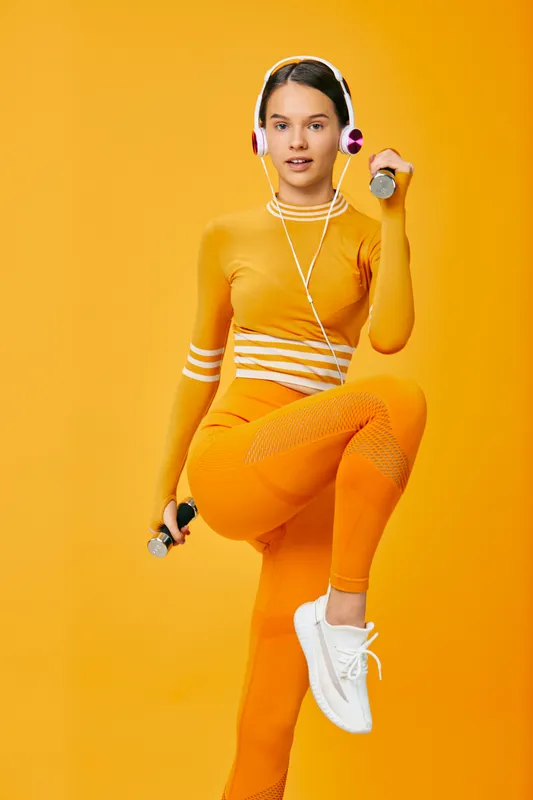 Young woman fitness portrait in bright orange activewear performing a high knee workout holding small dumbbells and wearing headphones against a solid orange background, energetic sport concept.