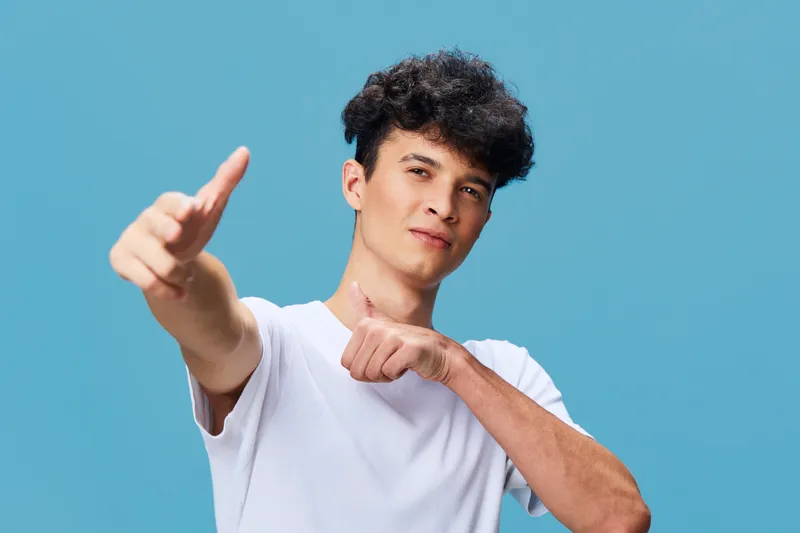 young man giving thumbs up gesture with confident expression wearing white t-shirt on blue background showing approval and positivity