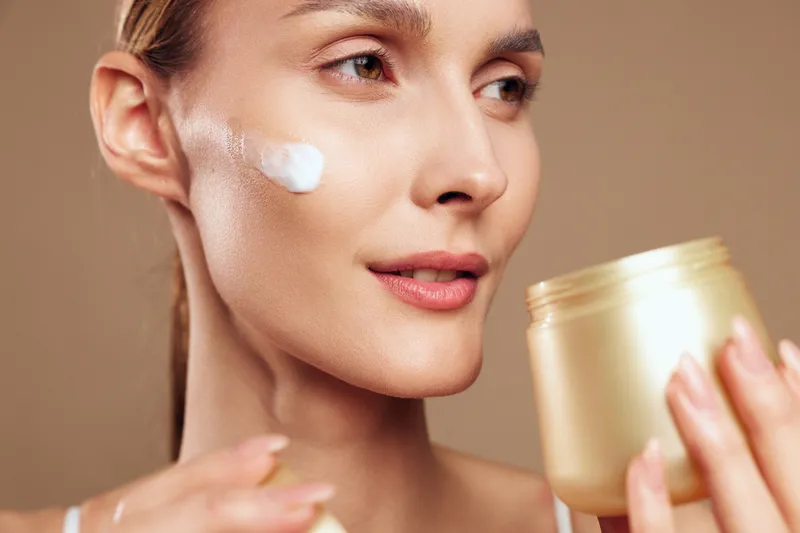 A closeup of a woman applying moisturizer to her cheek, a jar of cream visible in the foreground. Clear concept: skincare routine, hydration, and facial care.