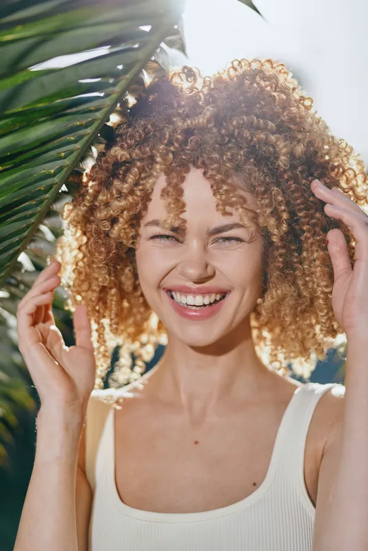 smiling woman with curly hair enjoying sunny day near tropical leaves, natural light, summer mood, joyful expression, casual white top outdoors