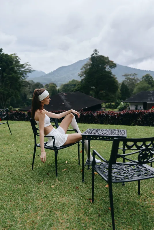 Young woman in a white outfit sitting on a black metal chair outdoors, displaying a calm emotion against a lush green mountain backdrop, perfect for summer aesthetics