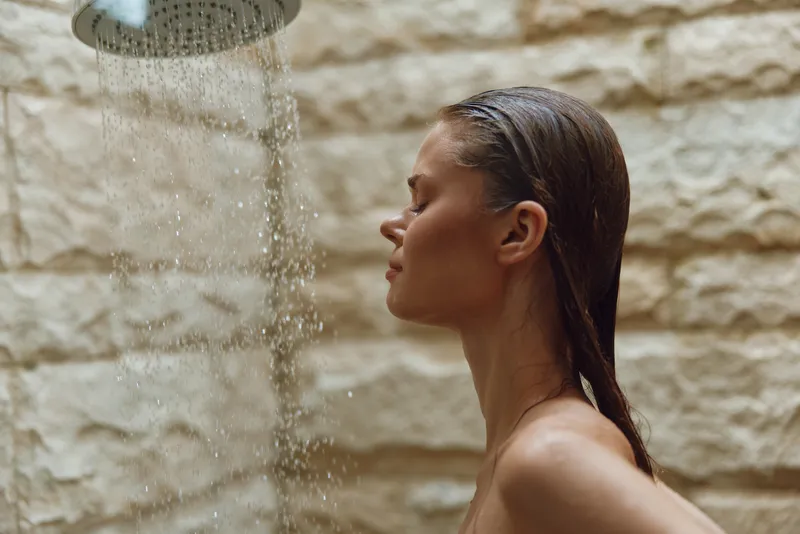 Relaxed woman under outdoor shower with wet hair, enjoying warm water and natural stone background in a peaceful moment of self-care and relaxation.