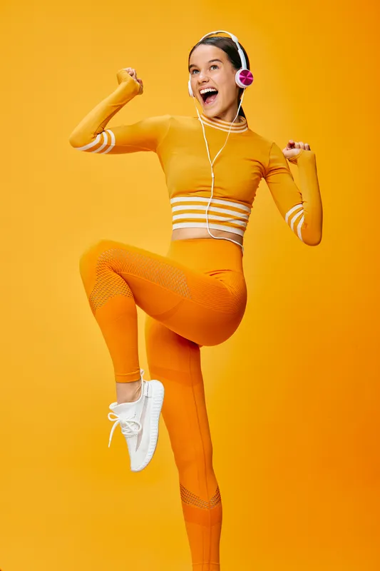 Woman fitness enthusiast wearing orange sportswear leaps with joy against a vivid orange background, headphones on, showcasing energy, style and active lifestyle.