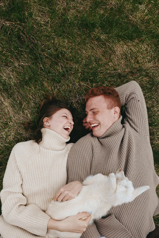 A smiling couple lounging on green grass in cozy sweaters, sharing a peaceful moment with a white cat between them. A lifestyle scene highlighting affection, comfort, and simple joy in an outdoor