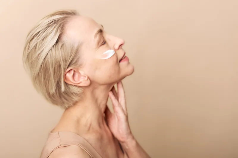 Profile of mature woman with short blonde hair applying cream on face, eyes closed and hand on neck. Concept of skincare, beauty, antiaging, and selfcare on beige background.