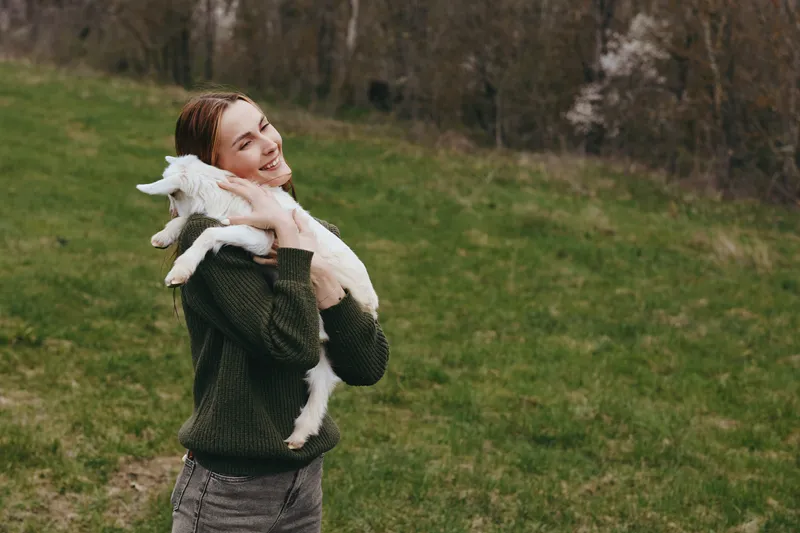 Joyful woman outdoors holds a goat kid in a grassy field, warm lifestyle moment, natural light, countryside scene, affectionate bond between human and animal, candid and wholesome today
