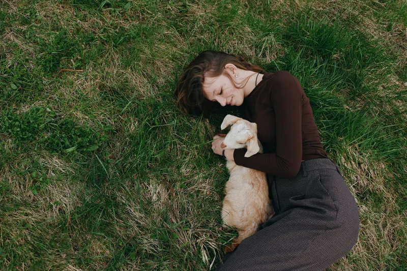 Young woman lying on grass cuddles a small goat in a peaceful outdoor setting, wearing a dark long-sleeve top and checkered pants, capturing a calm lifestyle moment.